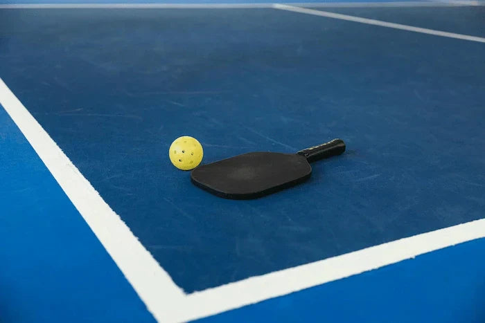 Raquette de pickleball et balle jaune posées sur un terrain bleu, comparaison pickleball et padel pour débutants