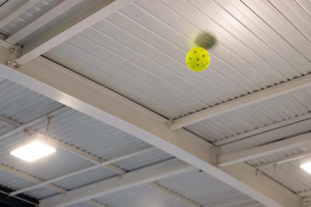 Balle de pickleball jaune en l'air, proche du plafond d'un terrain de pickleball intérieur.