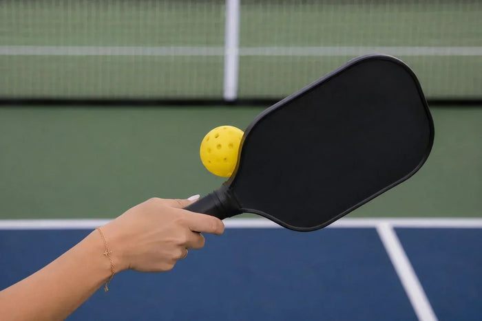 Main féminine tenant une raquette de pickleball lors de l’exécution d’un dink, avec une balle jaune en suspension au-dessus de la raquette sur un terrain de pickleball.
