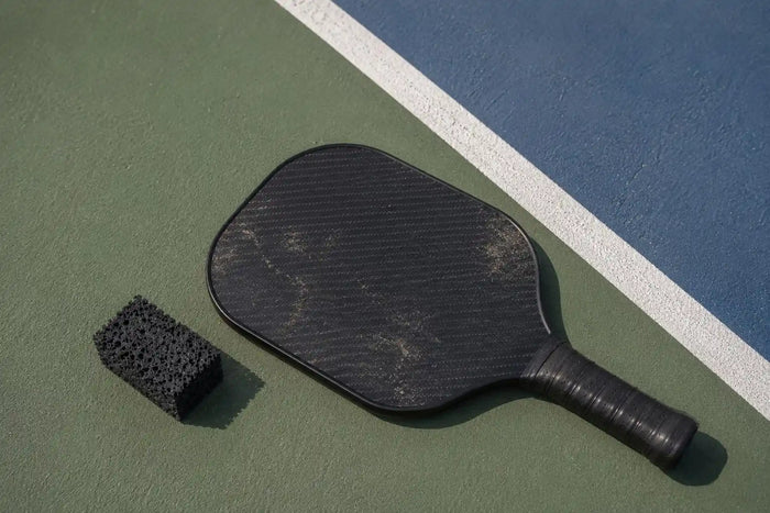 Raquette de pickleball en carbone posée au sol à côté d’une gomme de nettoyage sur un terrain extérieur.