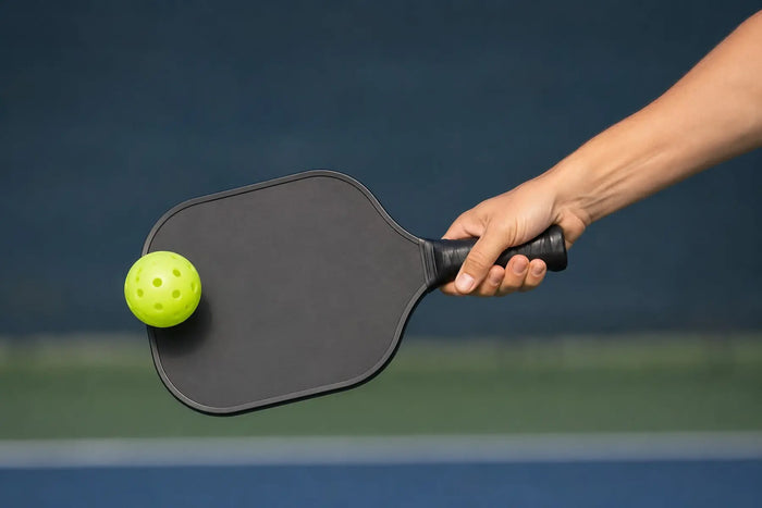 Gros plan d'une main frappant une balle de pickleball avec une raquette noire sur un terrain, illustrant une frappe pendant un échange.