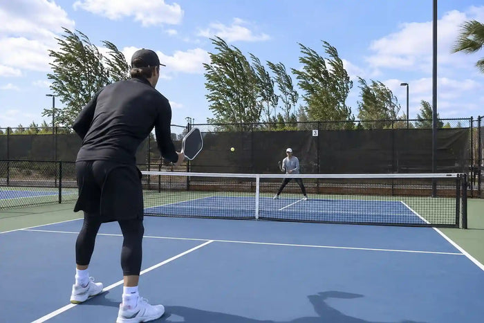 Deux joueurs disputant un échange sur un terrain de pickleball extérieur par temps venteux, avec des arbres pliés par le vent en arrière-plan.
