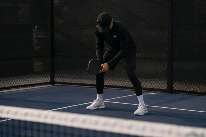 Joueur de pickleball en position d'attente près du filet avec raquette prête sur un terrain extérieur.