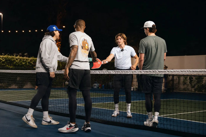 Quatre joueurs de pickleball se saluent au filet sur un terrain extérieur de nuit, portant une tenue de sport avec leggings de compression sous leurs shorts.