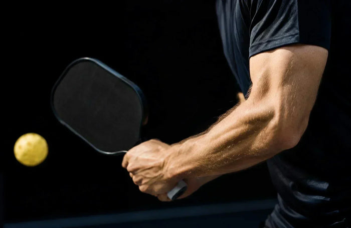 Bras d’un joueur de pickleball tenant une raquette noire avec balle en mouvement, mettant naturellement en avant la structure du coude et de l’avant-bras sur un terrain sombre.