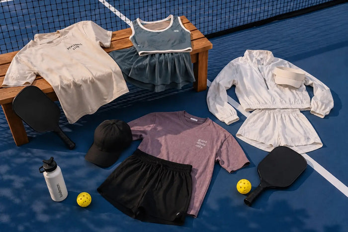 Différentes tenues de pickleball disposées sur un terrain, avec plusieurs ensembles en coton et en tissu technique, une casquette noire et des raquettes noires.