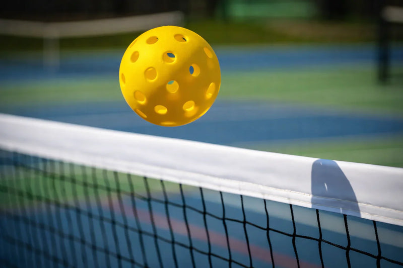 Balle de pickleball en plein vol au-dessus d'un filet de sport en extérieur.