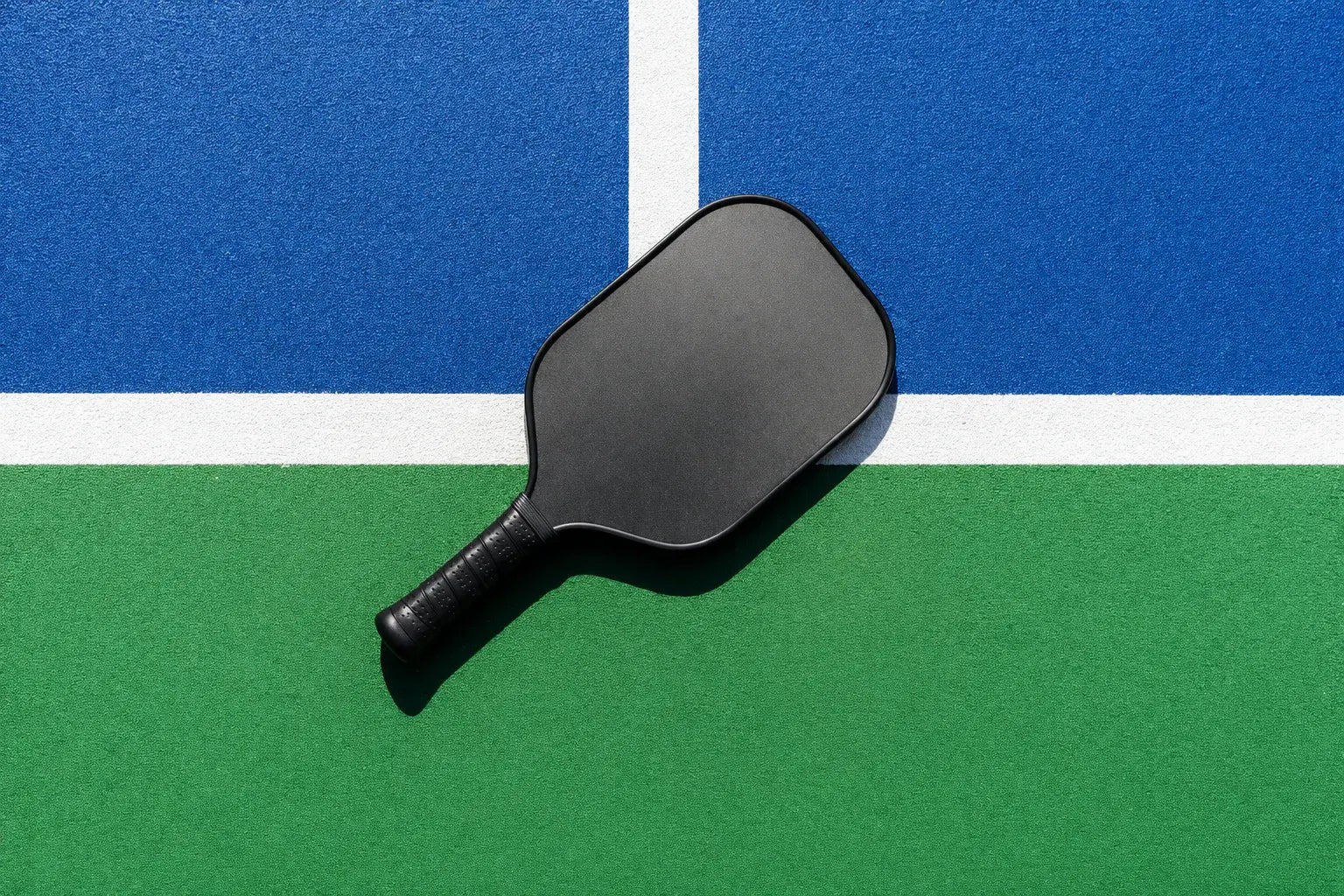 Raquette de pickleball noir posée sur la ligne d'un terrain bleu et vert