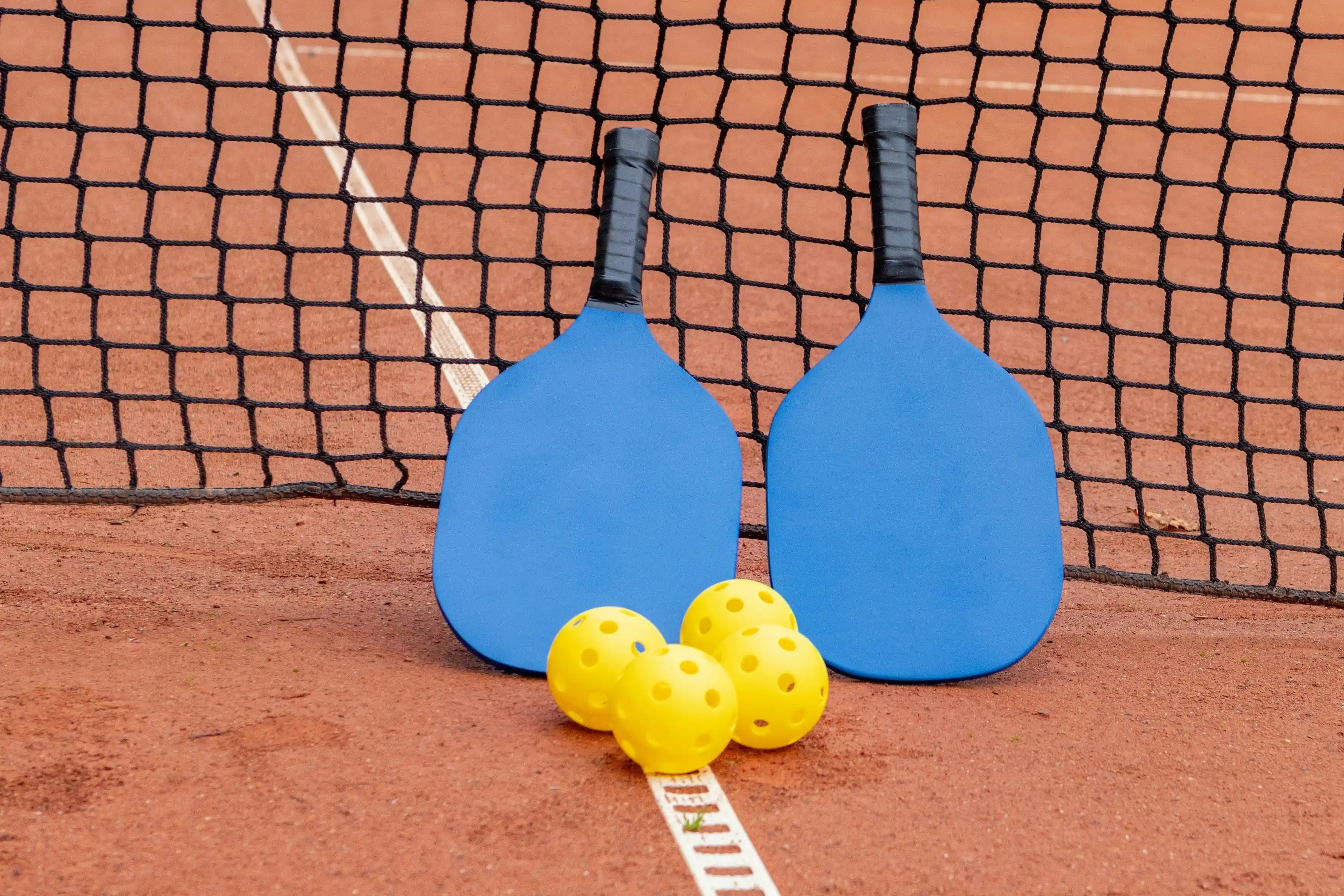Kit de pickleball comprenant deux raquettes bleues et quatre balles jaunes sur un terrain extérieur.