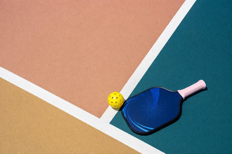 Raquette de pickleball bleue et balle jaune sur un terrain aux couleurs orange et bleu.