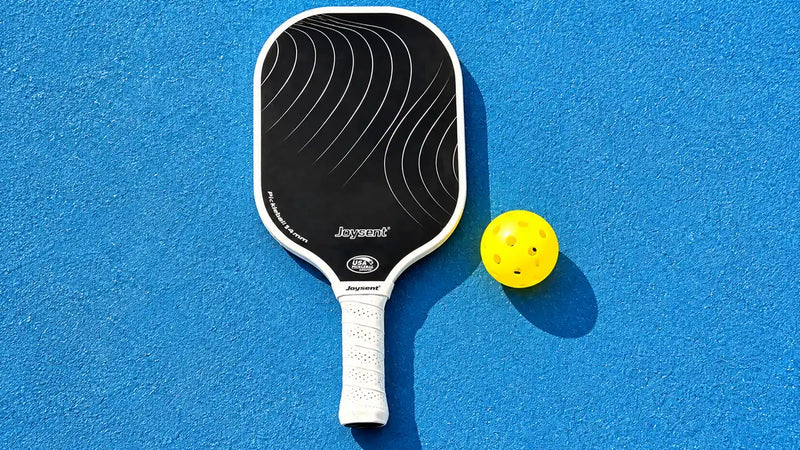 Raquette de pickleball Joysent noire et blanche de 14 mm posée sur un terrain bleu avec une balle jaune.