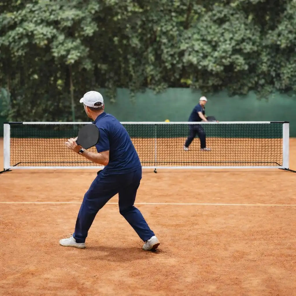 Match de pickleball en extérieur montrant le filet de 6,70 m installé sur un terrain en terre battue avec des joueurs en action.