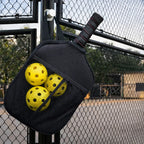 Housse de protection pour raquette de pickleball suspendue à un grillage de court, avec trois balles jaunes rangées dans la poche filet intégrée.