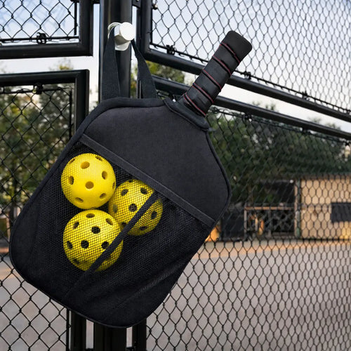 Housse de protection pour raquette de pickleball suspendue à un grillage de court, avec trois balles jaunes rangées dans la poche filet intégrée.
