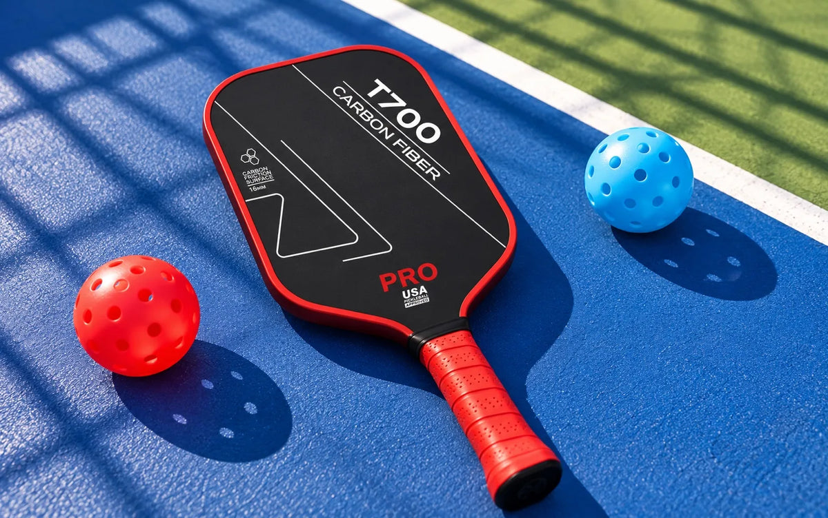 Raquette de pickleball T700 en carbone noir et rouge, accompagnée de deux balles rouge et bleue sur un terrain extérieur.